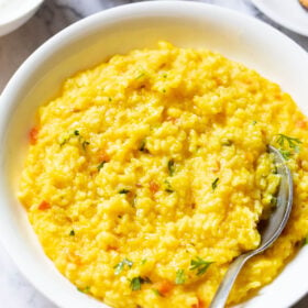 close up shot of dal khichdi in a bowl with a spoon inside with partial view of sides of curd, mango pickle in white bowls and roasted papad served in a white plate.