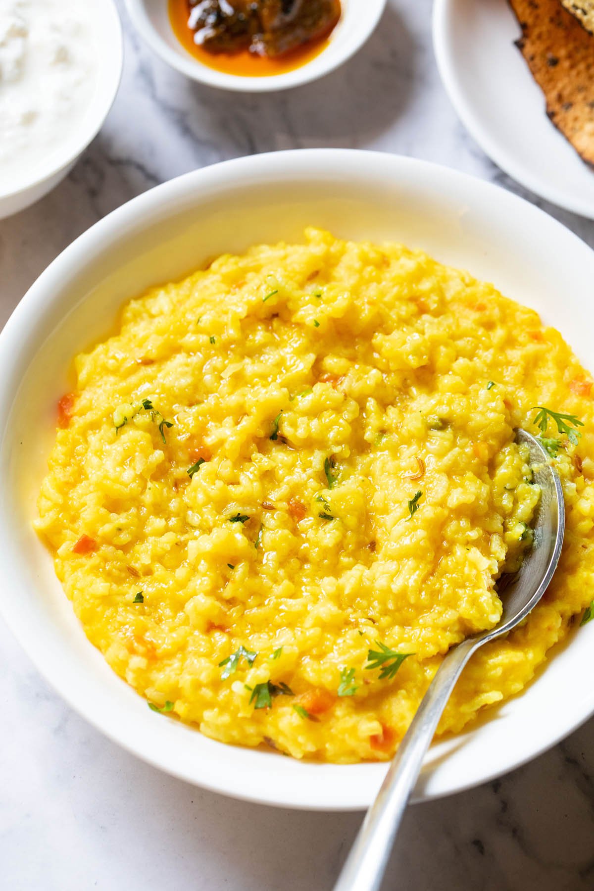 close up shot of dal khichdi in a bowl with a spoon inside with partial view of sides of curd, mango pickle in white bowls and roasted papad served in a white plate