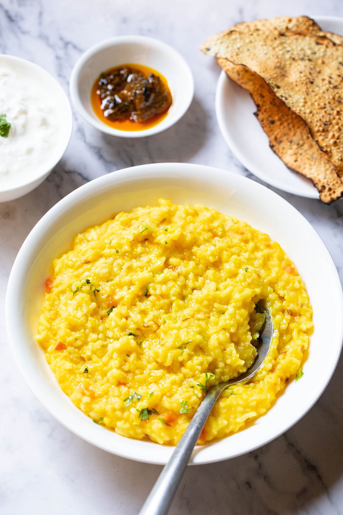 top shot of moong dal khichdi in a bowl with a spoon inside with sides of curd, mango pickle in white bowls and roasted papad served in a white plate