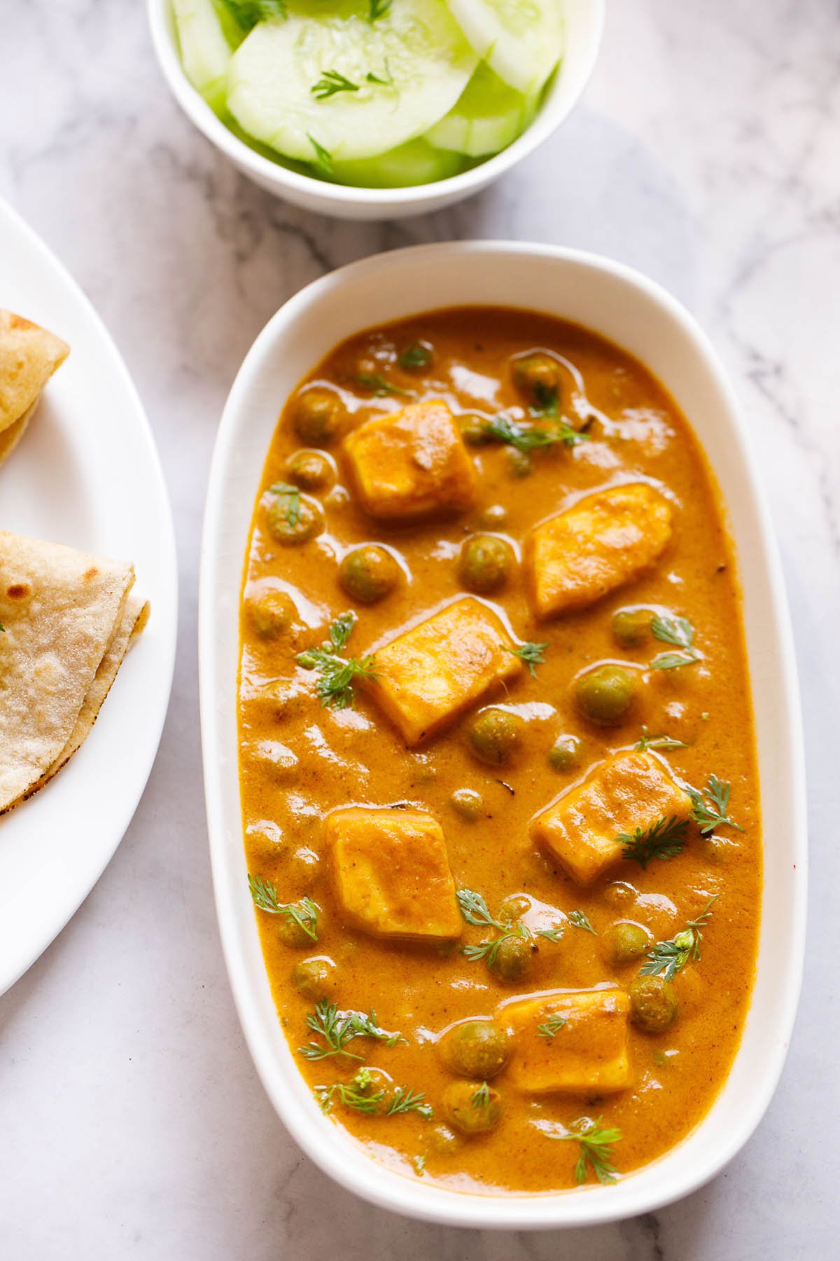 Top shot of matar paneer on white rectangle plate next to sliced cucumber in a white bowl and folded two roti in a white plate sitting on white marble table