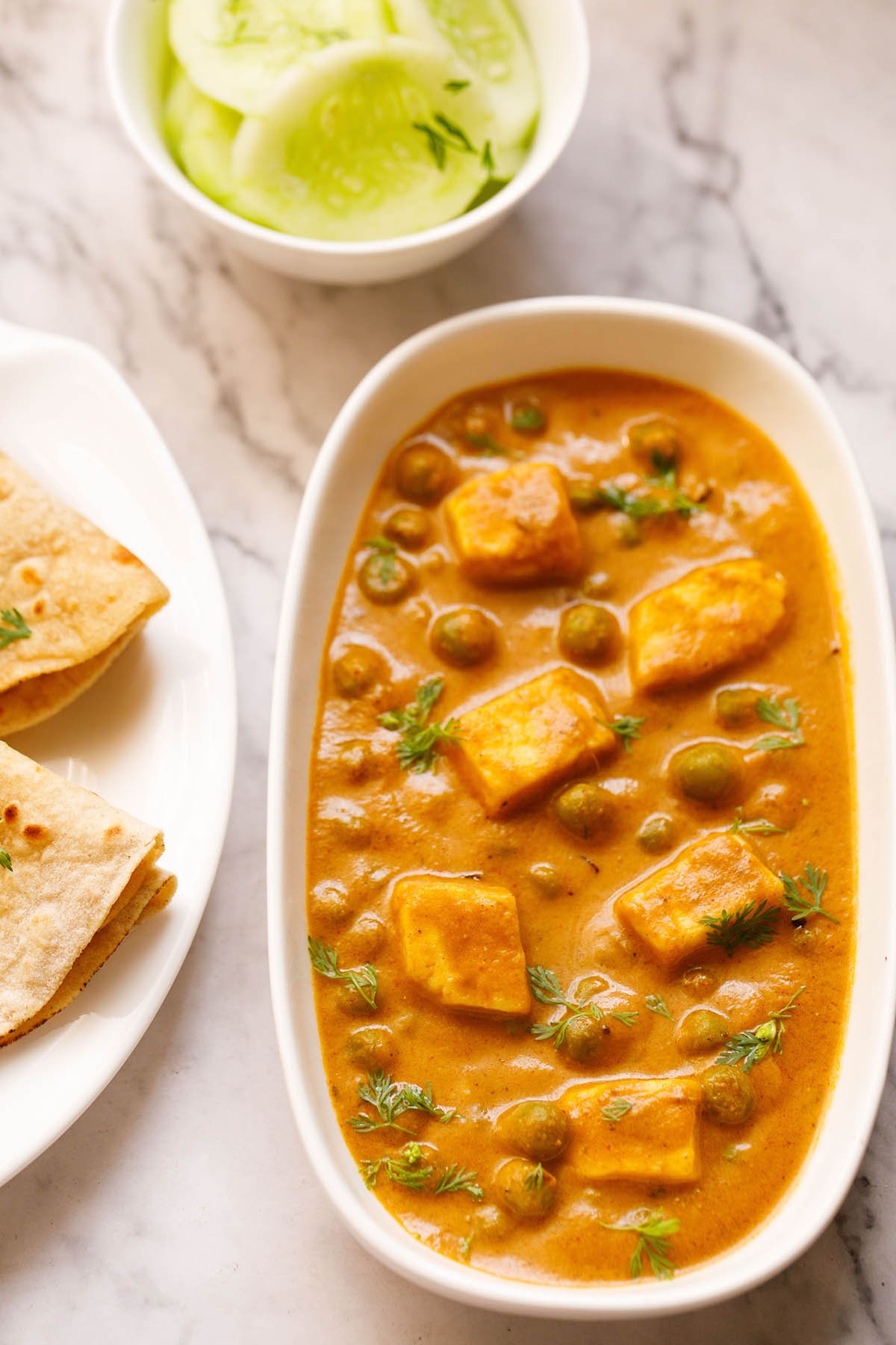 Top shot of matar paneer on white rectangle plate next to sliced cucumber in a white bowl and folded two roti in a white plate sitting on white marble table