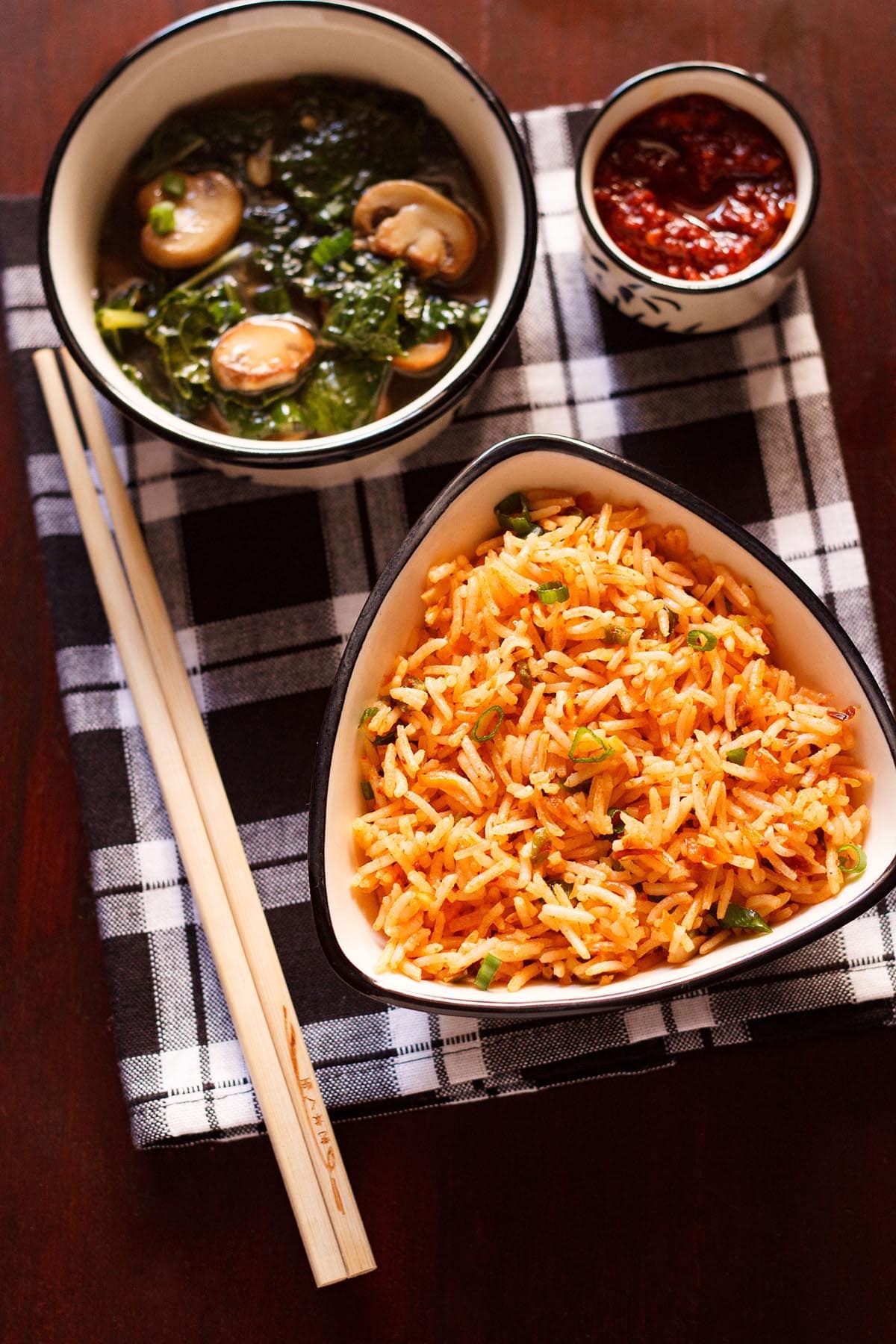 top shot of schezwan fried rice in a black rimmed triangular bowl with bamboo chopsticks at the side on a checkered black and white napkin