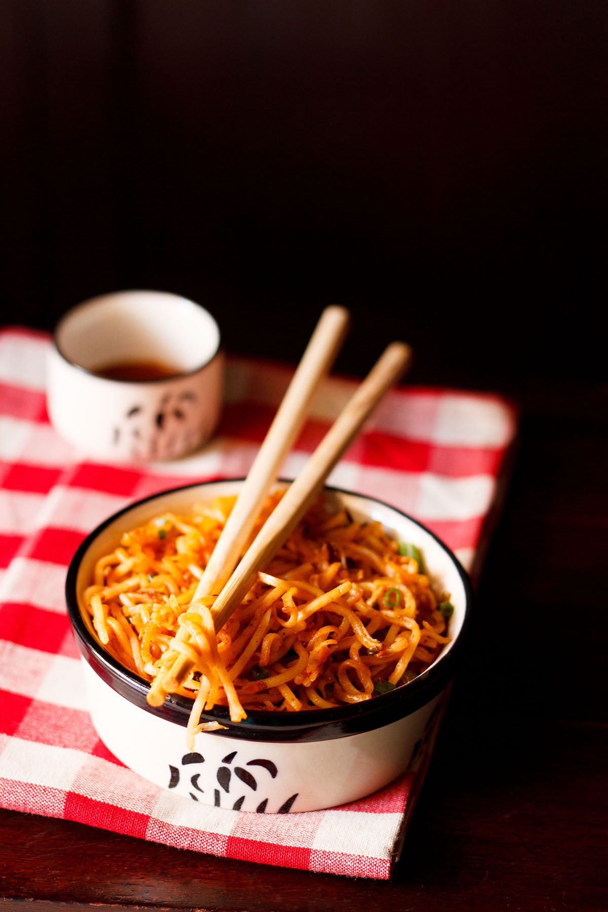 schezwan noodles in a black and cream bowl with a few noodle strands held between bamboo chopsticks placed on top of bowl on a checkered red and white napkin