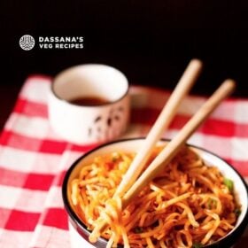 schezwan noodles in a black and cream bowl with a few noodle strands held between bamboo chopsticks placed on top of bowl on a checkered red and white napkin