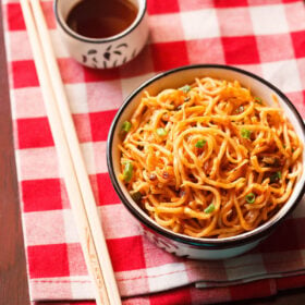 schezwan noodles in a black rimmed cream bowl with bamboo chopsticks at the side and a small bowl of schezwan sauce placed on a checkered red and white napkin