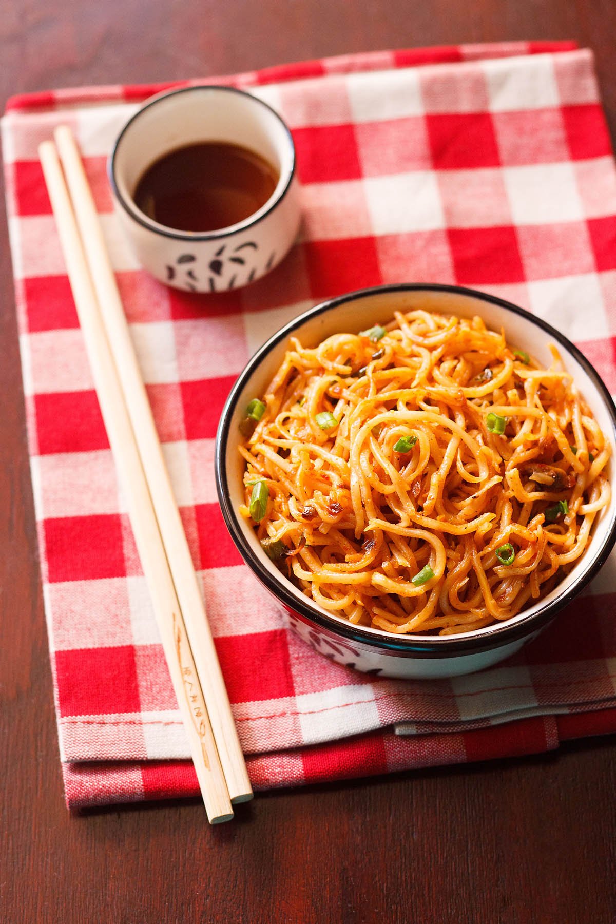 schezwan noodles in a black rimmed cream bowl with bamboo chopsticks at the side and a small bowl of schezwan sauce placed on a checkered red and white napkin