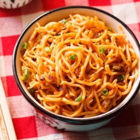 schezwan noodles in a black rimmed cream bowl with bamboo chopsticks at the side on a checkered red and white napkin