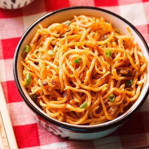 schezwan noodles in a black rimmed cream bowl with bamboo chopsticks at the side on a checkered red and white napkin