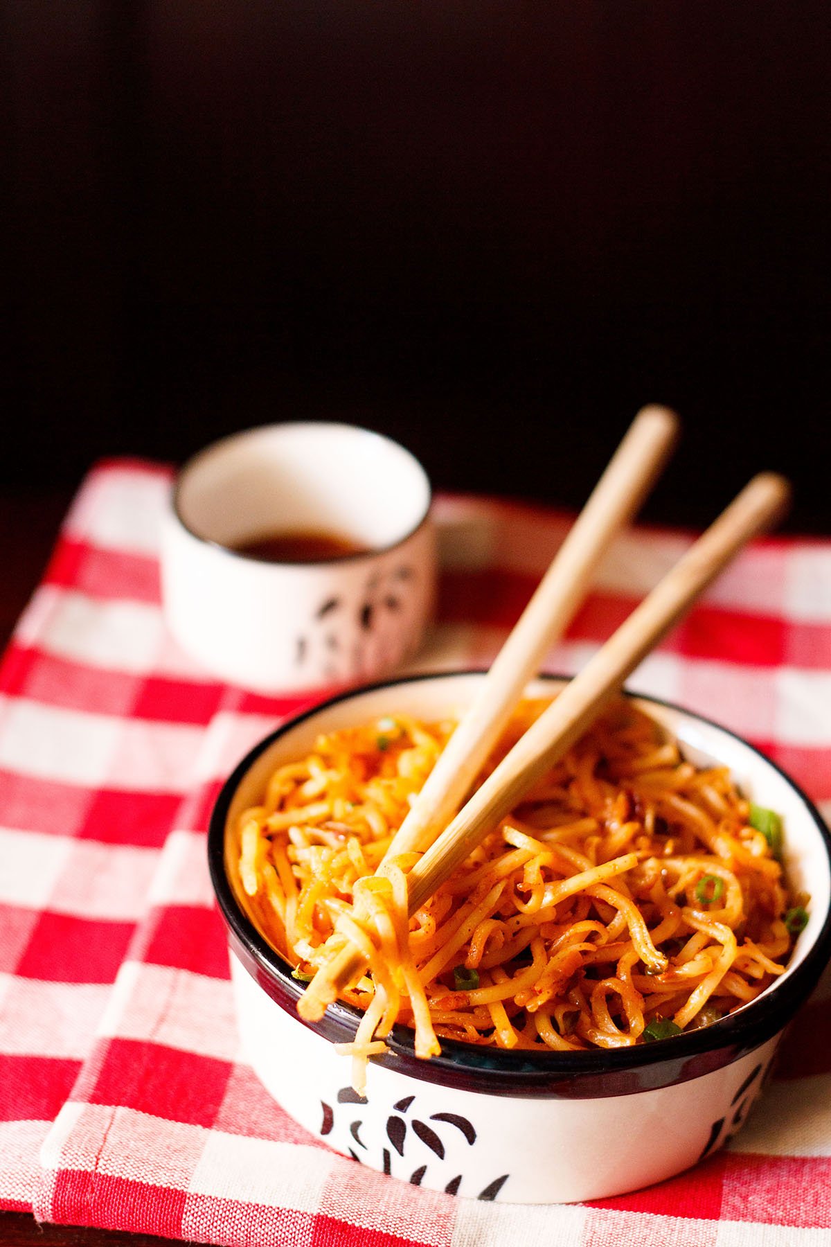 schezwan noodles in a black and cream bowl with a few noodle strands held between bamboo chopsticks placed on top of bowl on a checkered red and white napkin