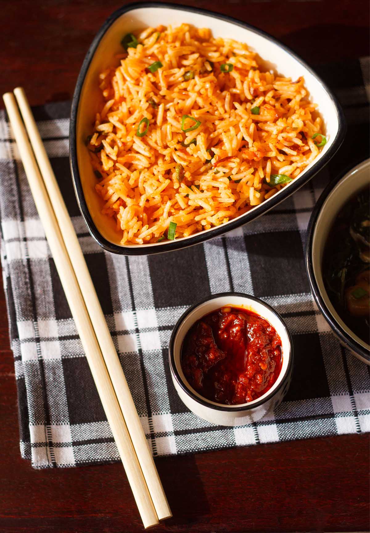 overhead shot of schezwan sauce in a small bowl with chopsticks and schezwan fried rice on a black and white checkered napkin