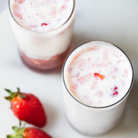 top photo of two glasses of strawberry milk with two strawberries placed at the side on a white marble board