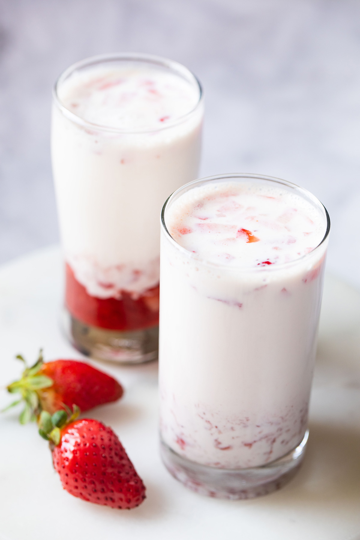 two collins glasses with homemade strawberry milk and two fresh strawberries on a white surface