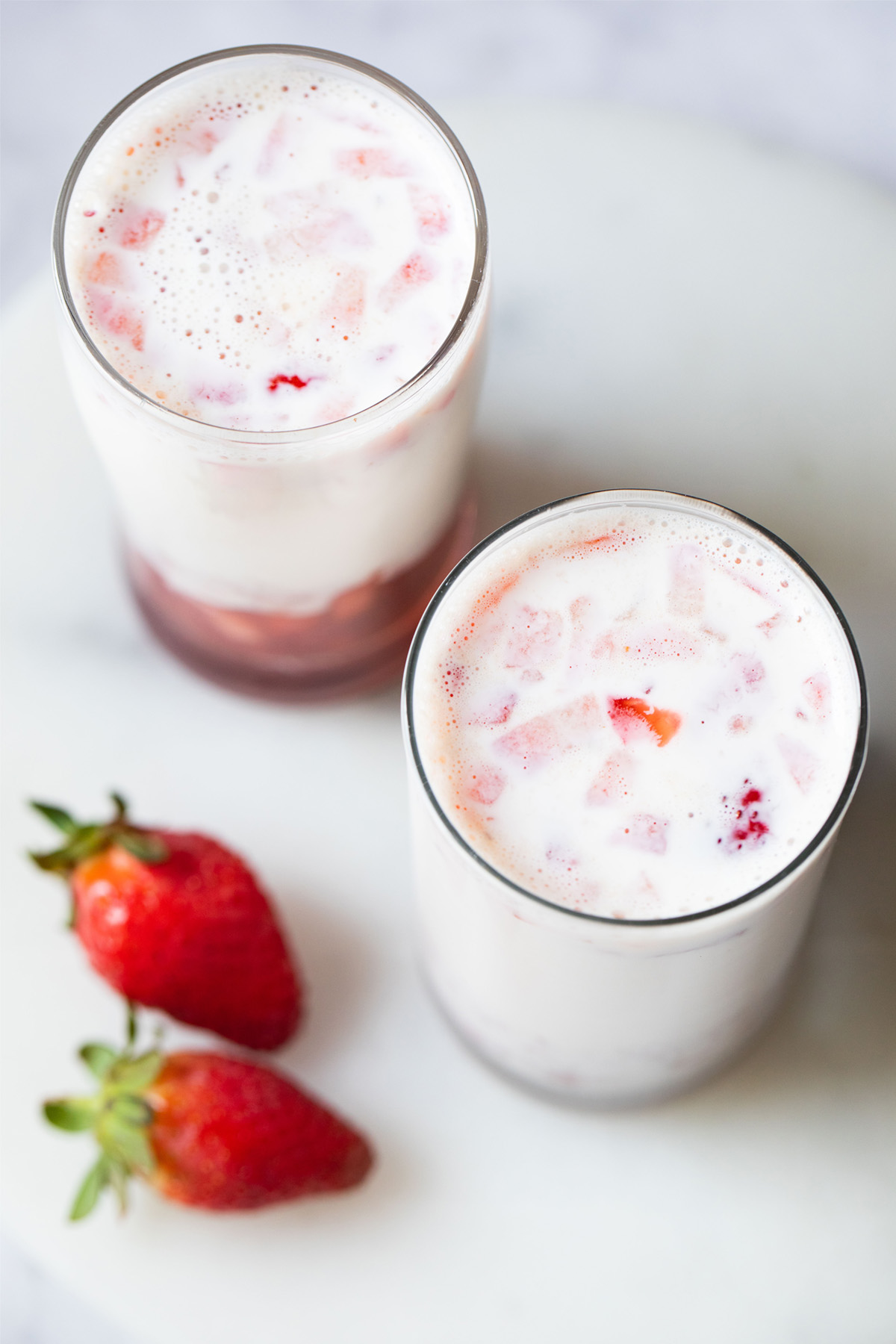 top photo of two glasses of strawberry milk with two strawberries placed at the side on a white marble board