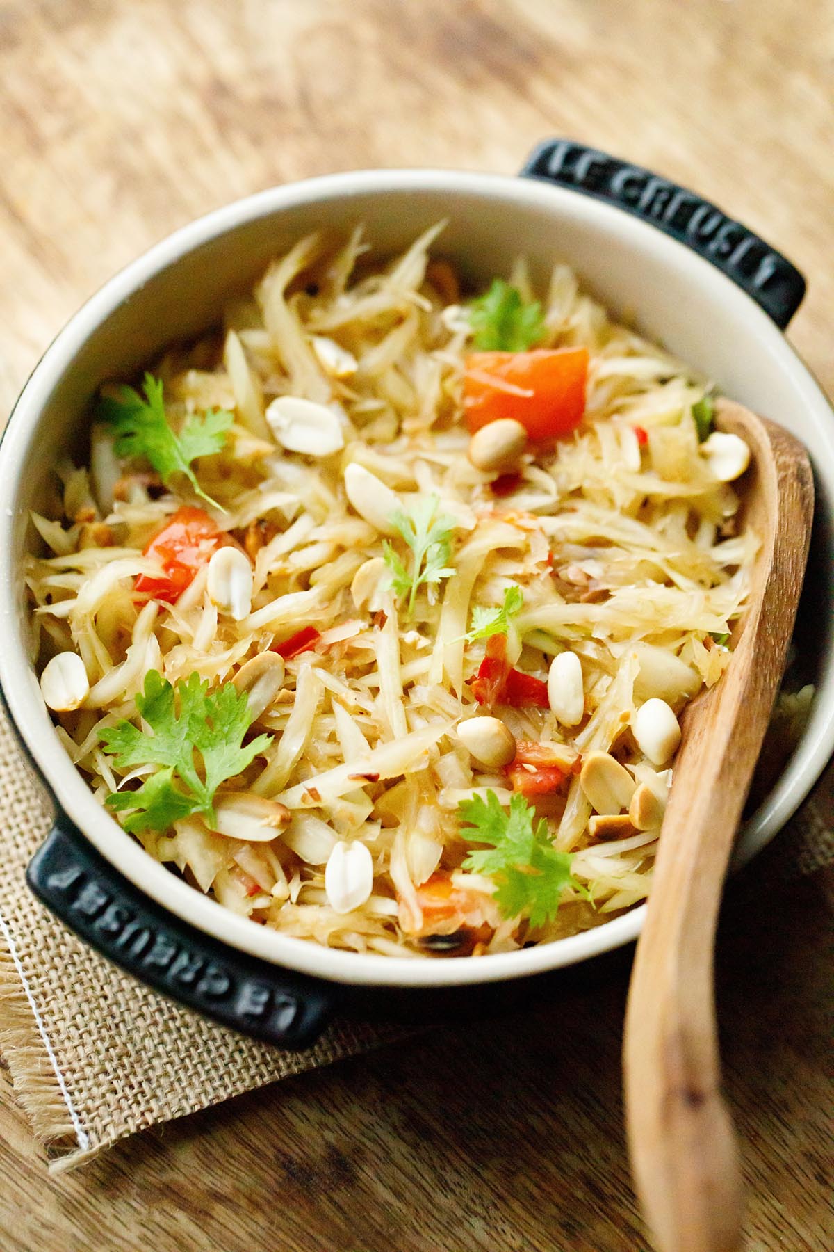 thai green papaya salad garnished with fresh cilantro and roasted peanuts in a black le creuset dish with a wooden spoon