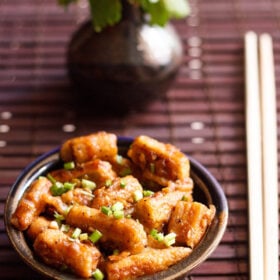 chilli baby corn garnished with spring onion greens in a dark brown ceramic bowl with bamboo chopsticks on right side on a dark brown bamboo mat