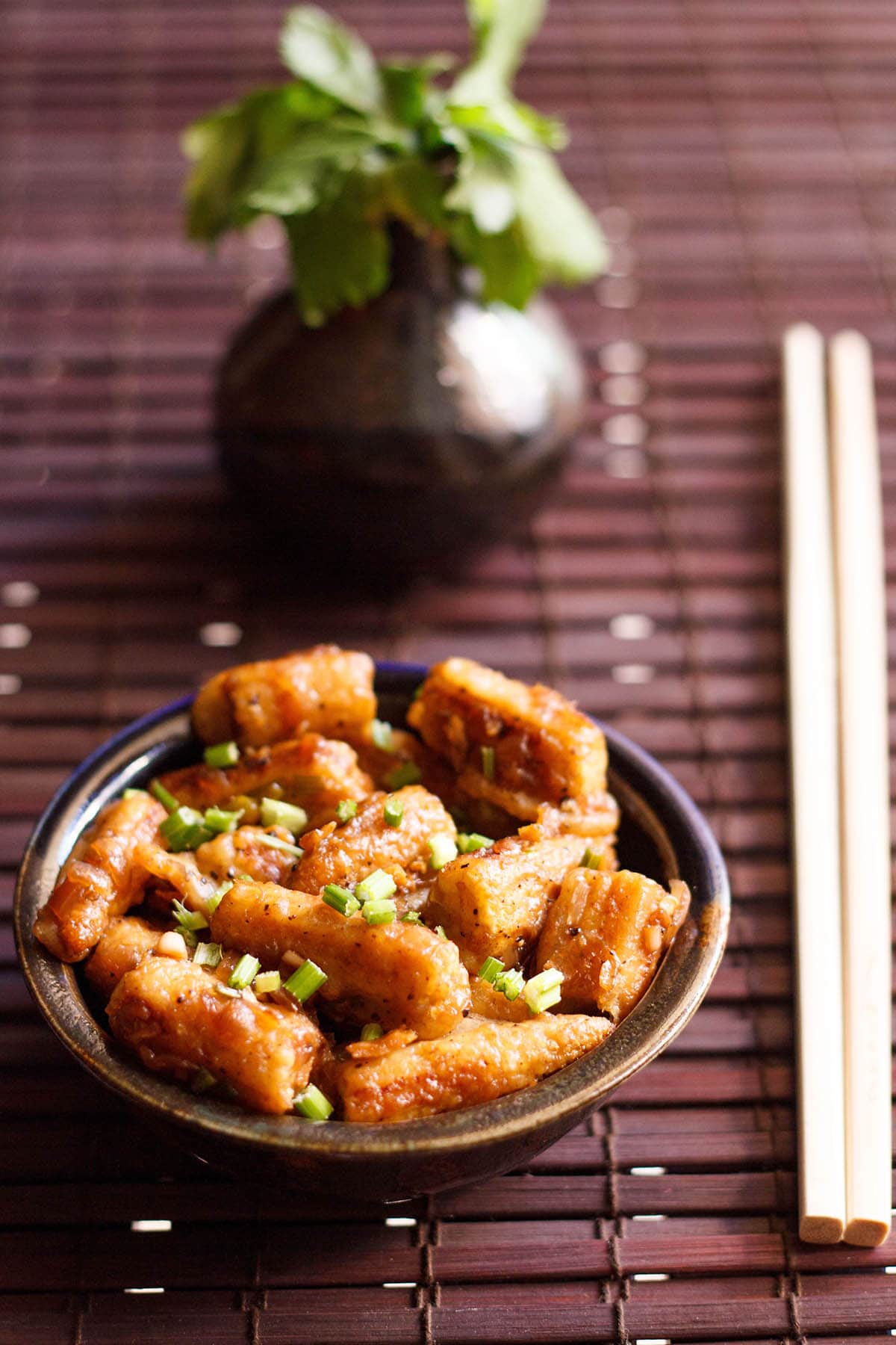 chilli baby corn garnished with spring onion greens in a dark brown ceramic bowl with bamboo chopsticks on right side on a dark brown bamboo mat