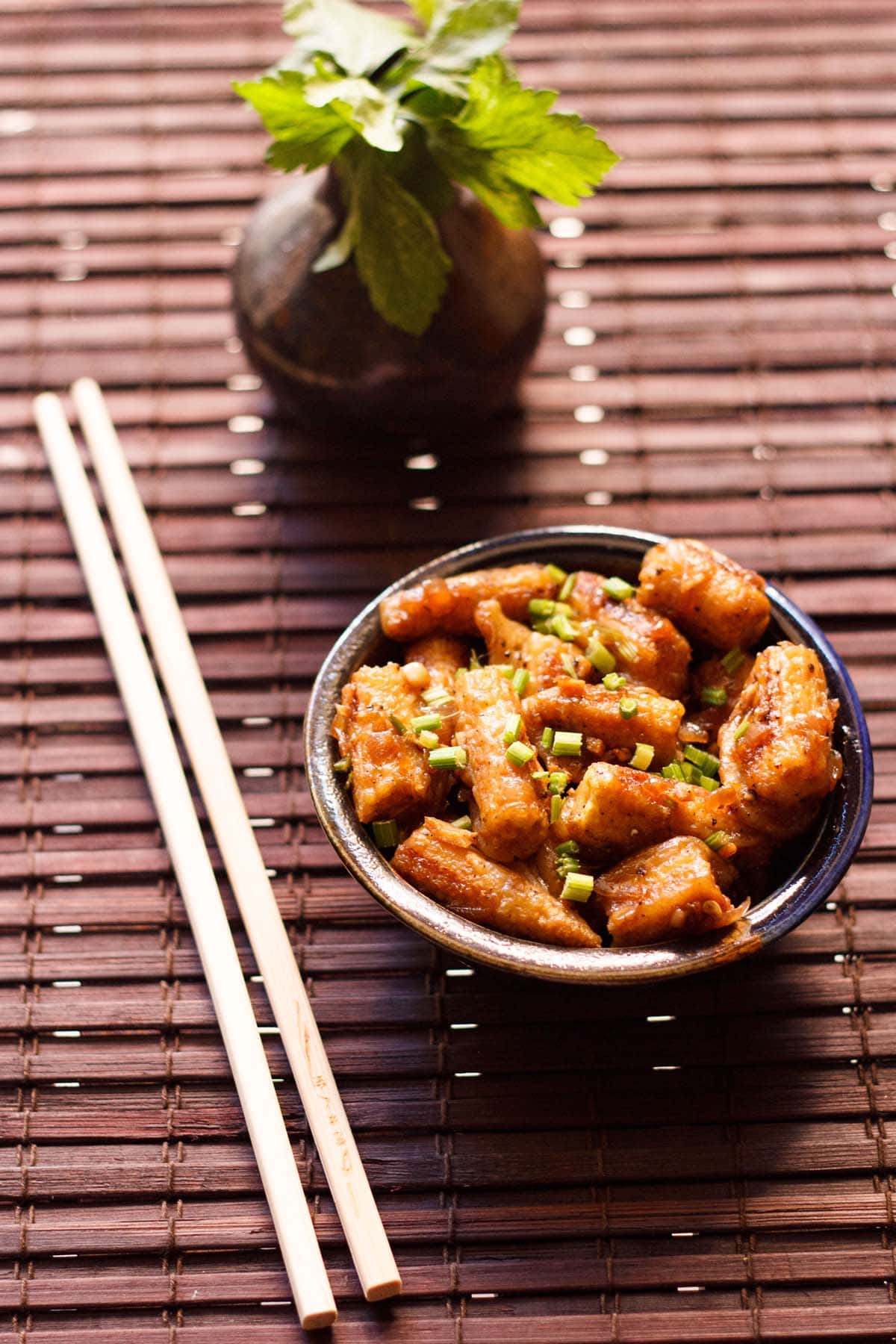 chilli baby corn garnished with spring onion greens in a dark brown ceramic bowl with bamboo chopsticks on left side on a dark brown bamboo mat