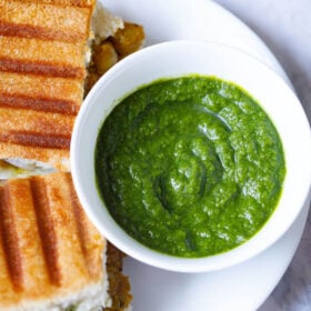 Top shot of coriander chutney in white bowl next to grilled sandwich on white plate