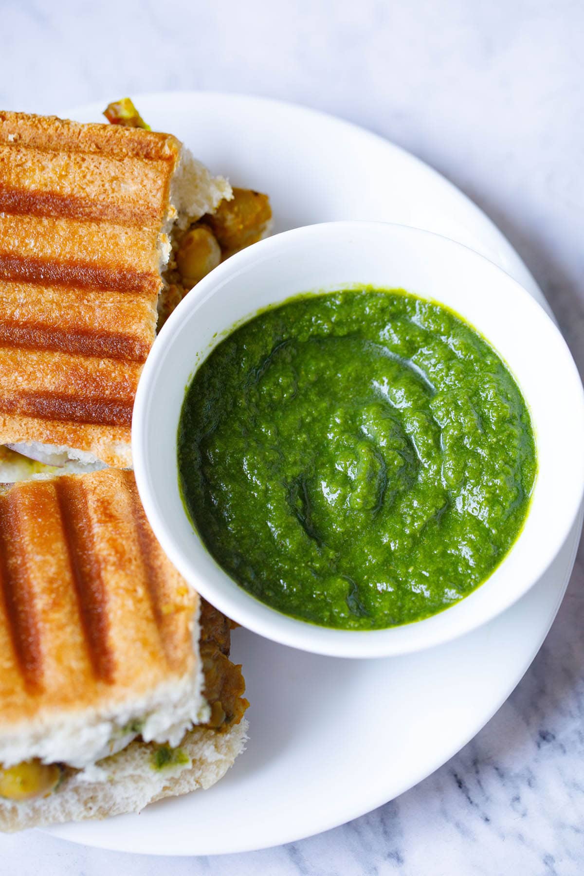 top shot of coriander chutney in white bowl placed next to grilled sandwiches on a white plate