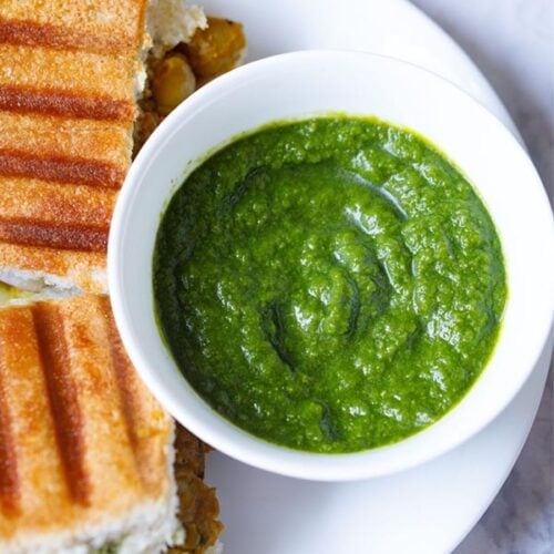 top shot of cilantro chutney in white bowl placed next to grilled sandwiches on a white plate