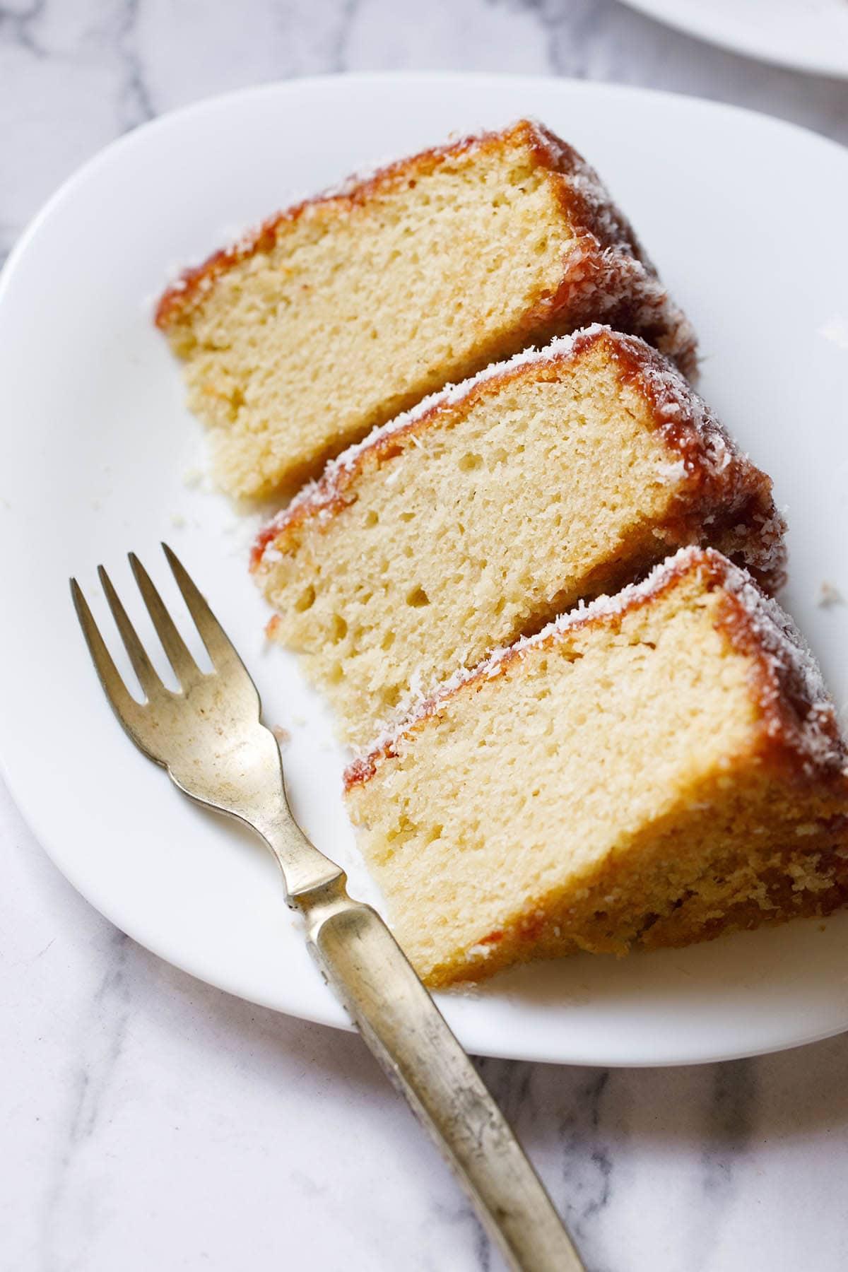 close up shot of thick slices of eggless vanilla cake on a square shaped white plate woth a brass fork on the plate