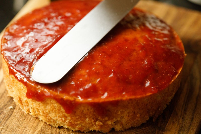 spreading strawberry jam glaze on eggless vanilla cake with an offset spatula