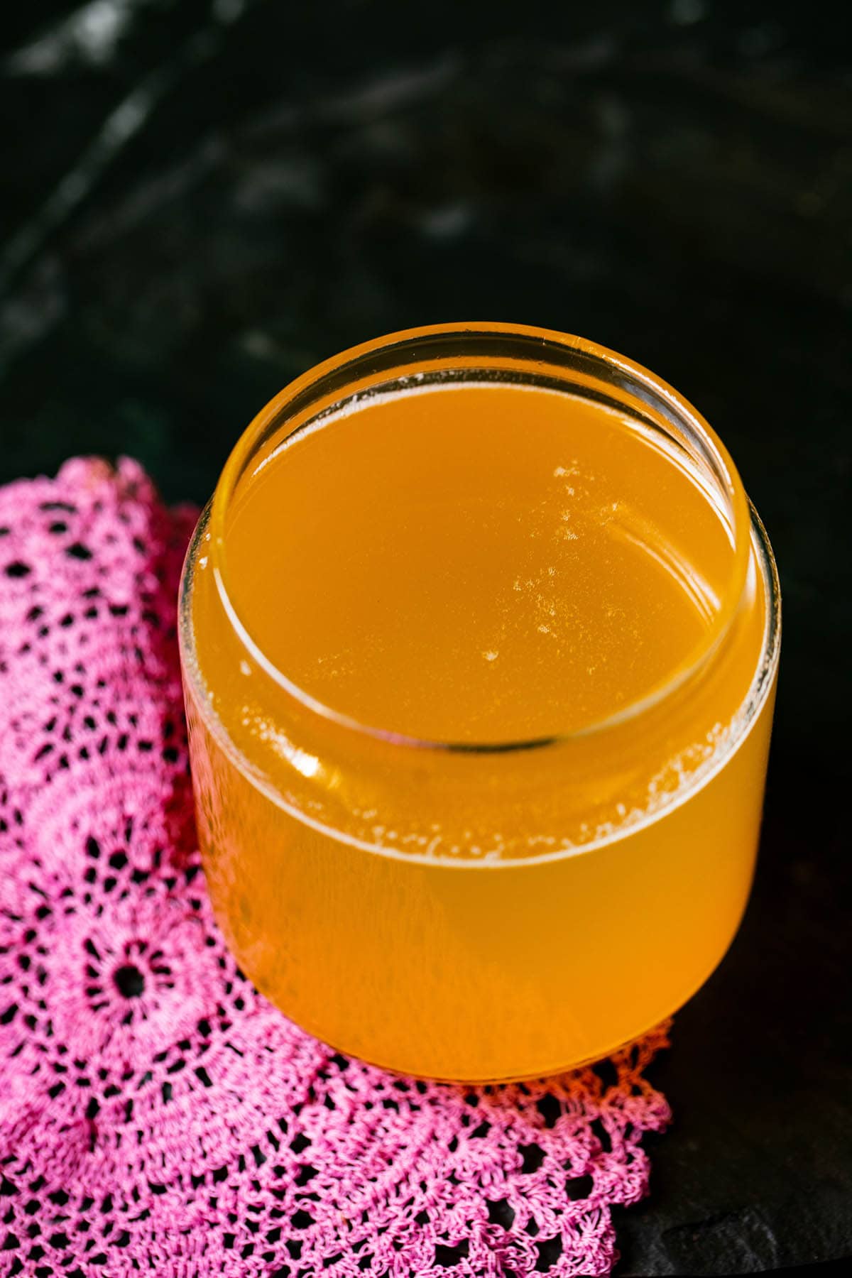 clear liquid ghee in a glass jar with a dark pink doily by the side on a black surface