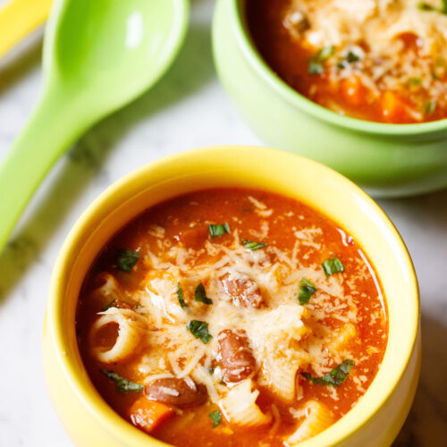 overhead shot of vegetable minestrone soup in a yellow bowl garnished with parmesan cheese and fresh basil