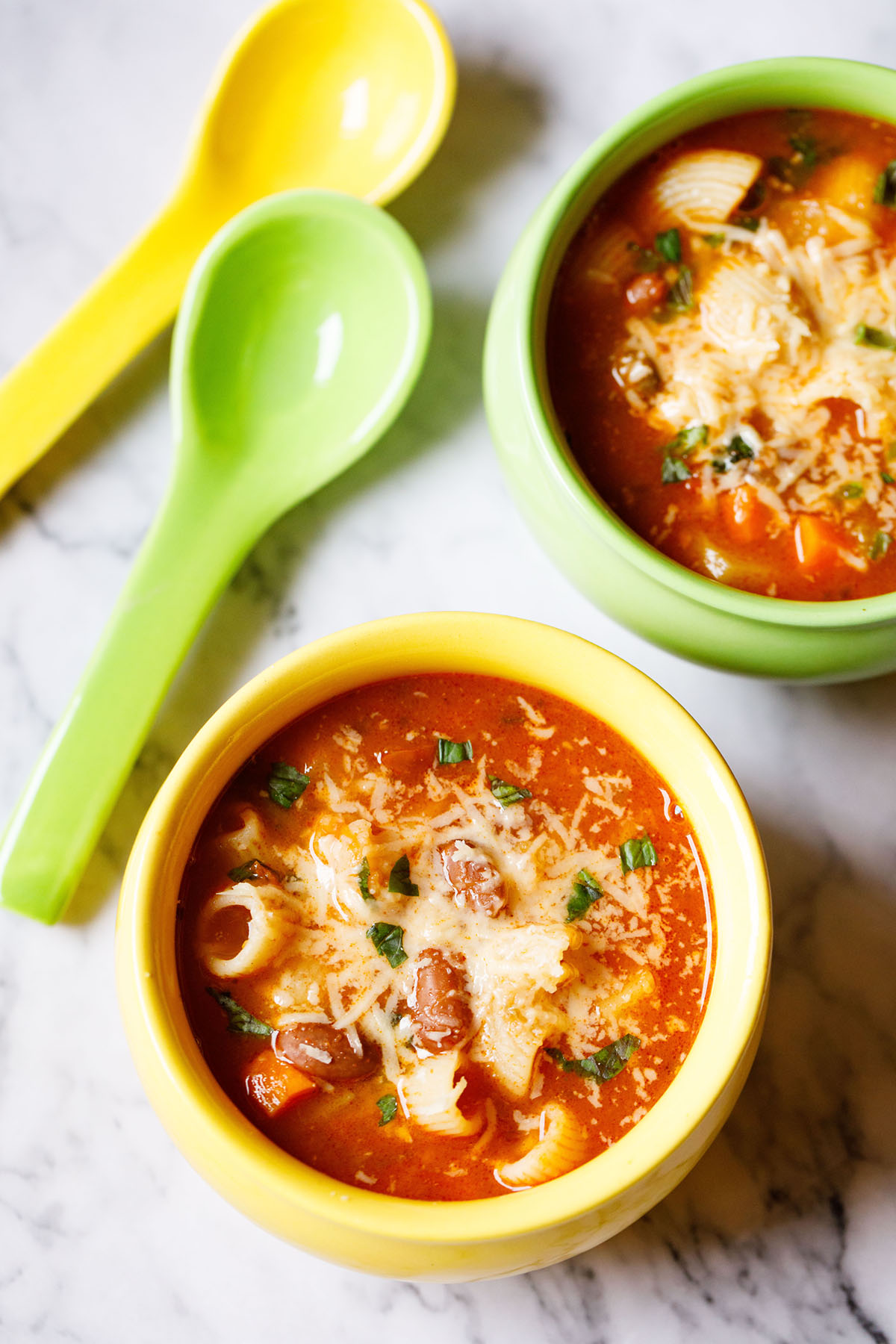 one light green and one light yellow enamel bowls filled with vegetable minestrone soup, garnished with parmesan cheese and basil with matching enamel spoons