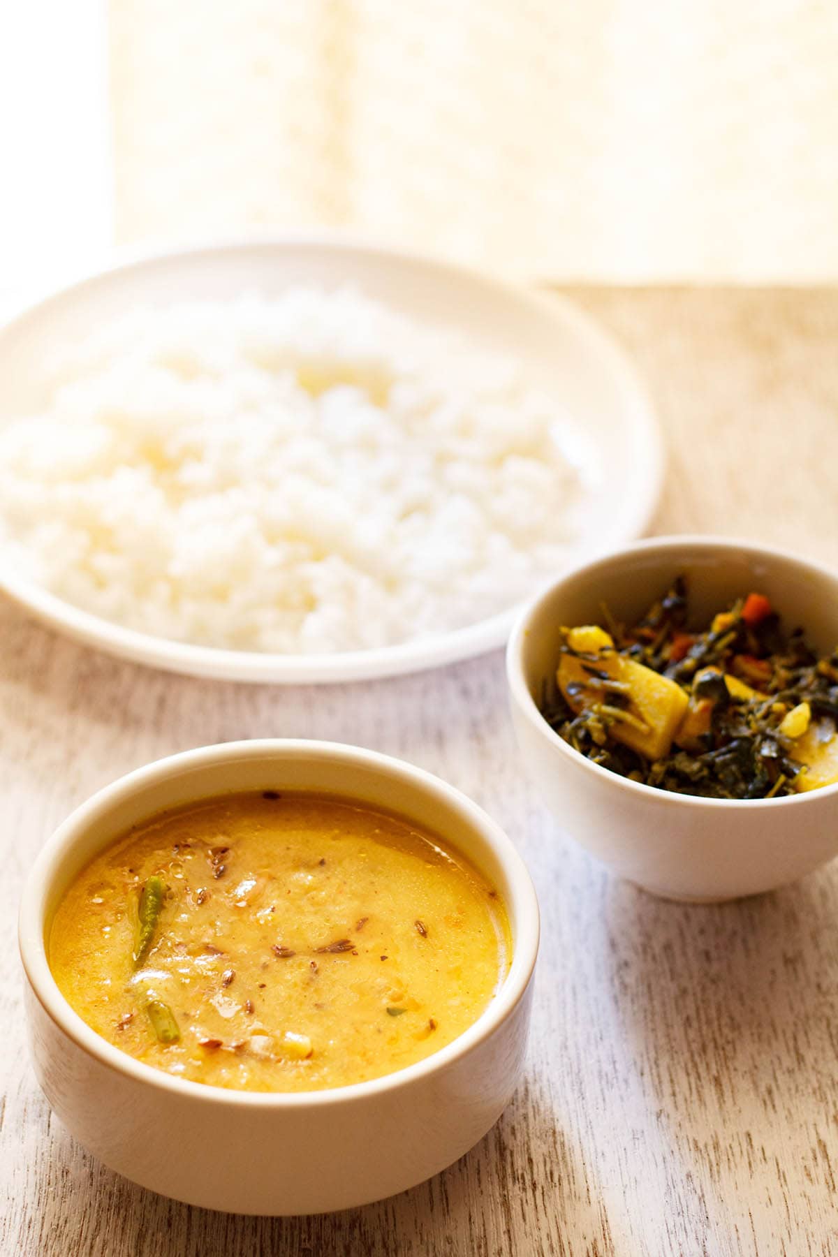 moong dal recipe in white bowl with a plate of steamed rice and a vegetable dish in the background