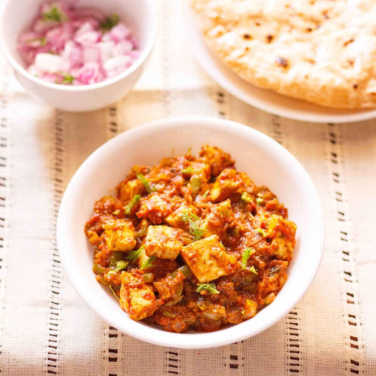 paneer tawa masala with coriander leaves garnish in a white bowl on a cream linen with a side plate of roti and a small side bowl of chopped red onions and coriander