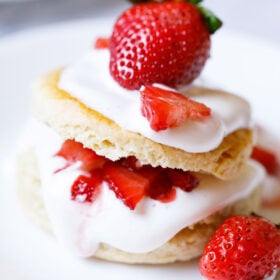 close up shot of strawberry shortcake with the cream and strawberry filling seen and topped with some and a large strawberry on top on a white plate