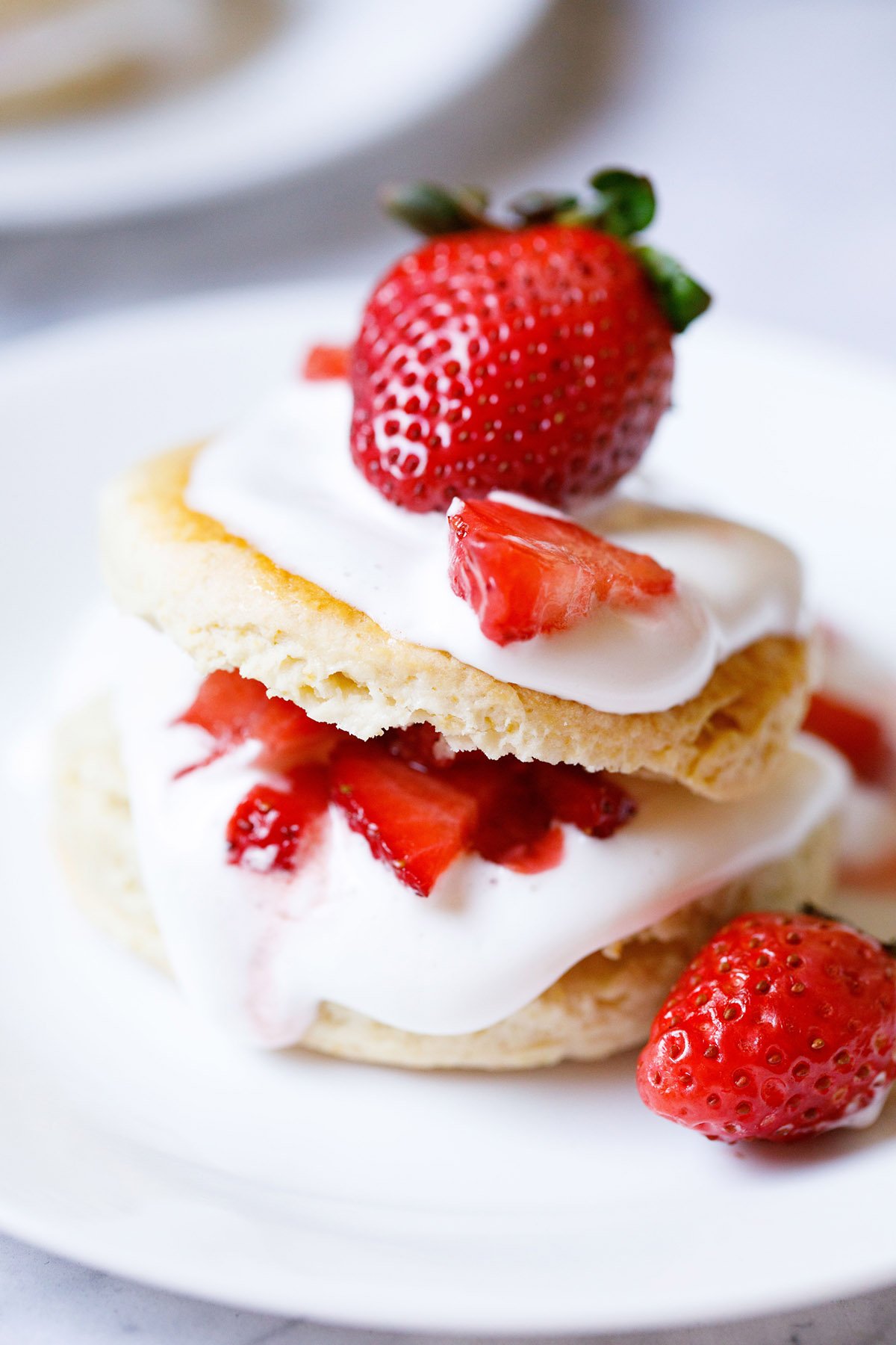 close up shot of strawberry shortcake with the cream and strawberry filling seen and topped with some and a large strawberry on top on a white plate