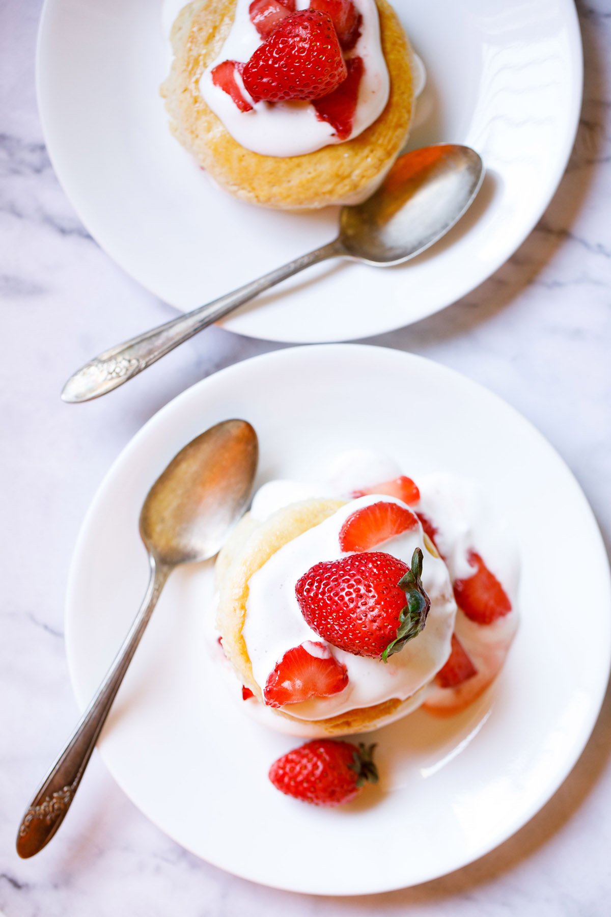 top shot of strawberry shortcakes topped with cream and strawberries each on two white plates with a brass spoon on each plate on white marble background