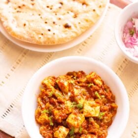 overhead shot of tawa paneer with coriander leaves garnish in a white bowl on a cream linen with a side plate of roti and a small side bowl of chopped red onions and coriander leaves