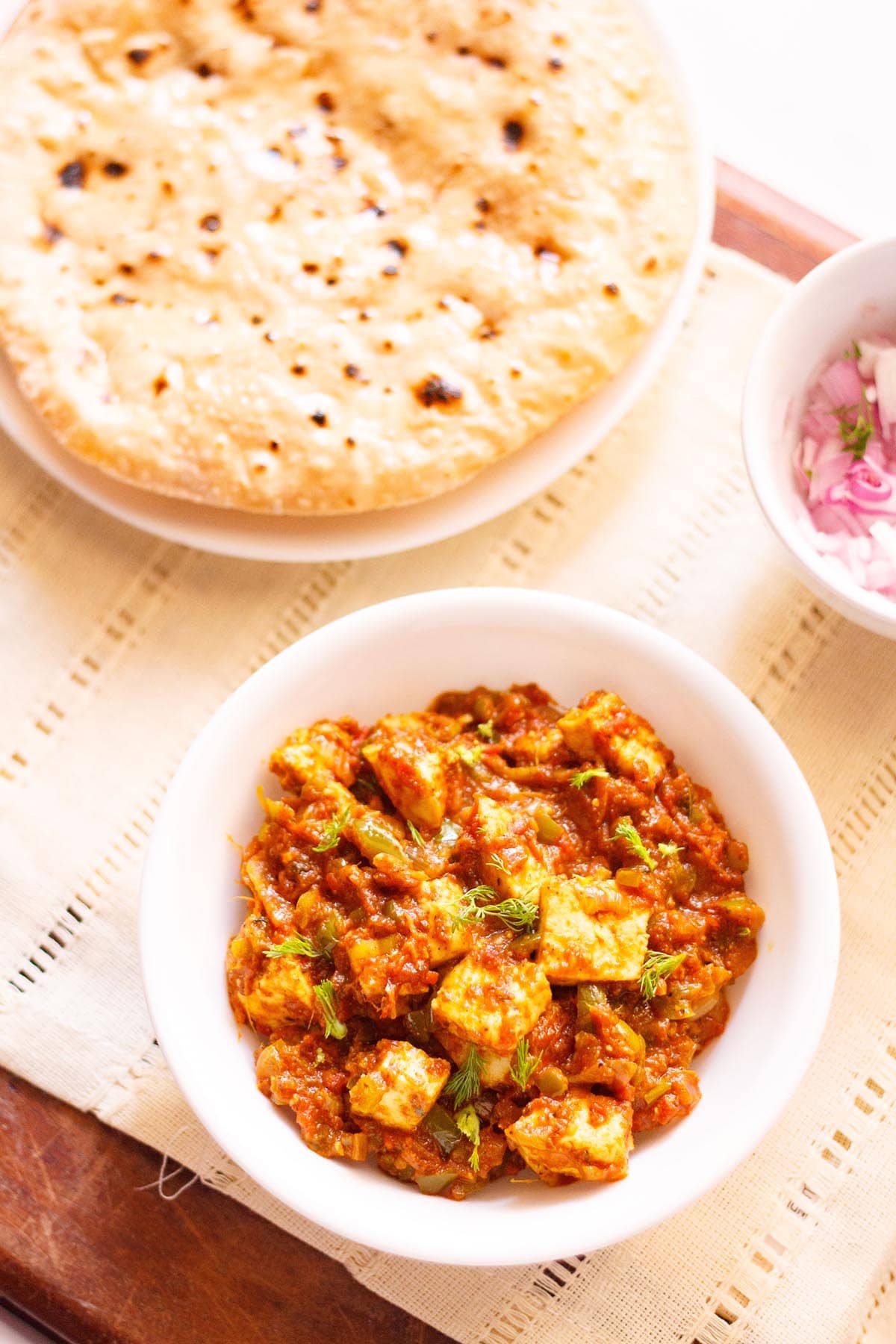 overhead shot of tawa paneer with coriander leaves garnish in a white bowl on a cream linen with a side plate of roti and a small side bowl of chopped red onions and coriander leaves