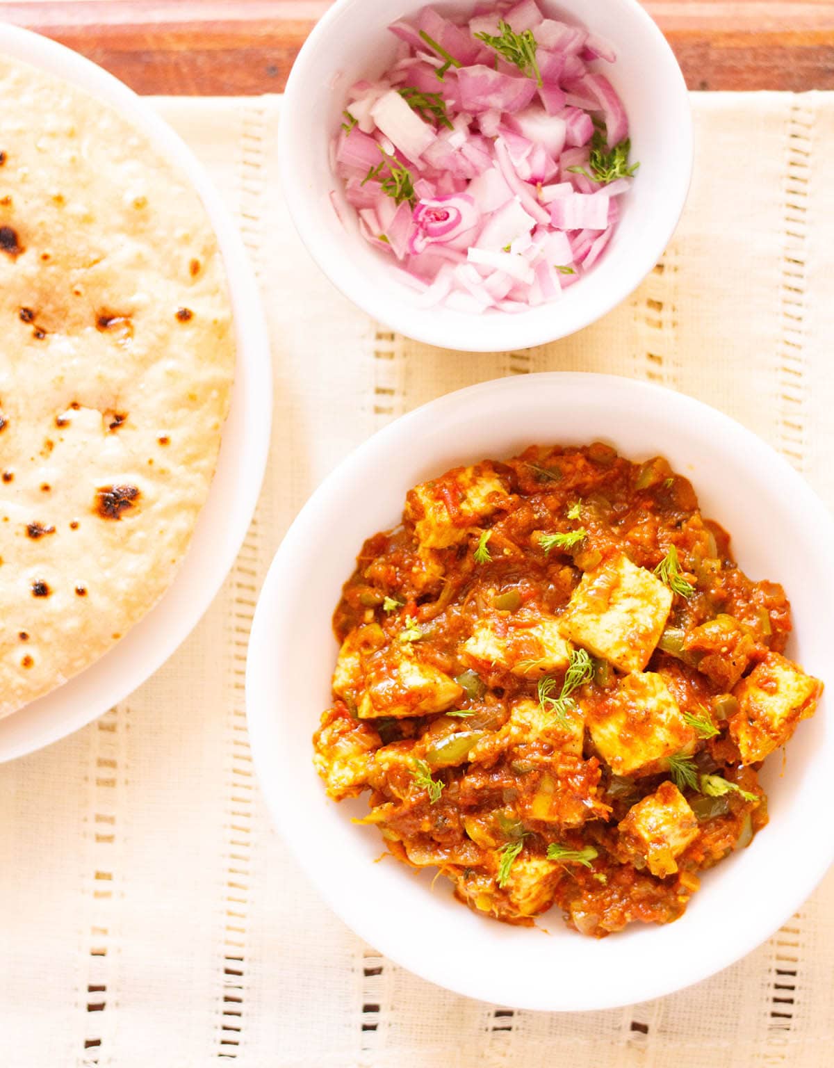 overhead shot of tawa paneer with coriander leaves garnish in a white bowl on a cream linen with a side plate of roti and a small side bowl of chopped red onions and coriander leaves