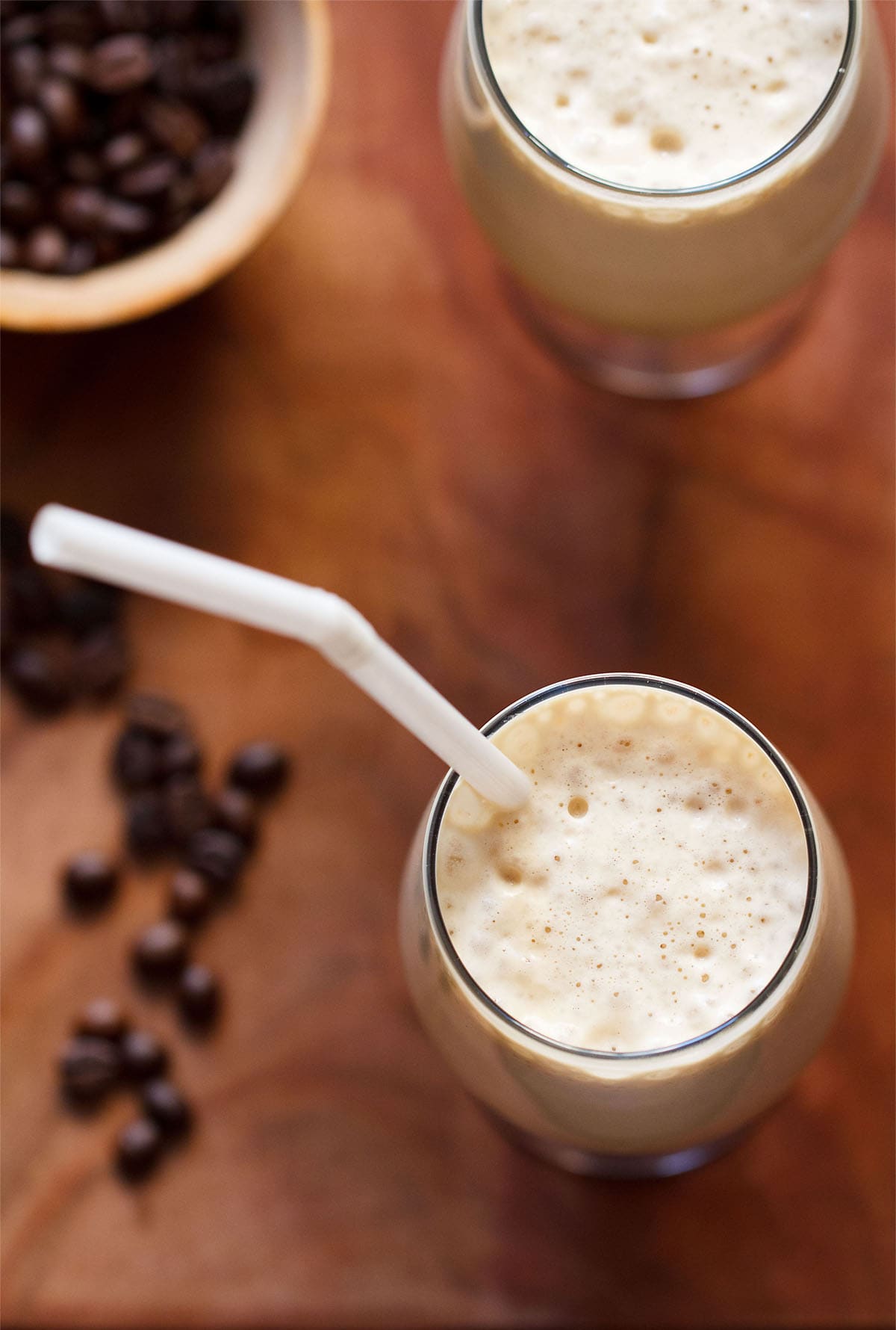 top shot of cold coffee served in two glasses with straw in one glass and coffee beans in a bowl and on a brown wooden board