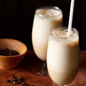 side shot of cold coffee in two glass with a white straw in one glass with coffee beans in a dark orange bowl and some coffee beans on a wooden board
