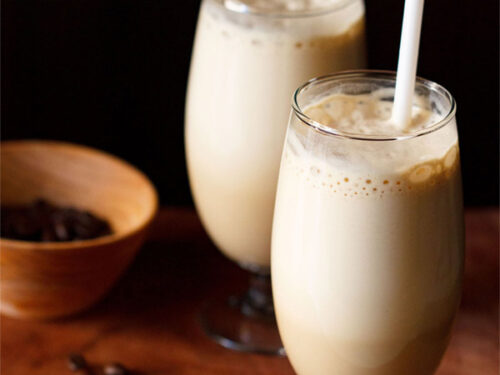 side shot of cold coffee in two glasses with a white straw in one glass with coffee beans in a dark orange bowl and some coffee beans on a wooden board