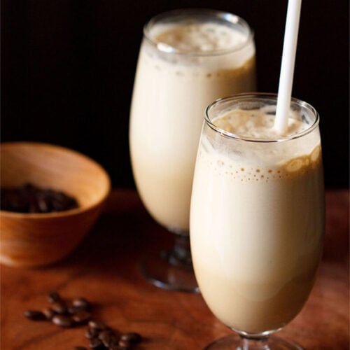 toma lateral de café frío en dos vasos con una pajita blanca en un vaso con granos de café en un tazón naranja oscuro y algunos granos de café en una tabla de madera