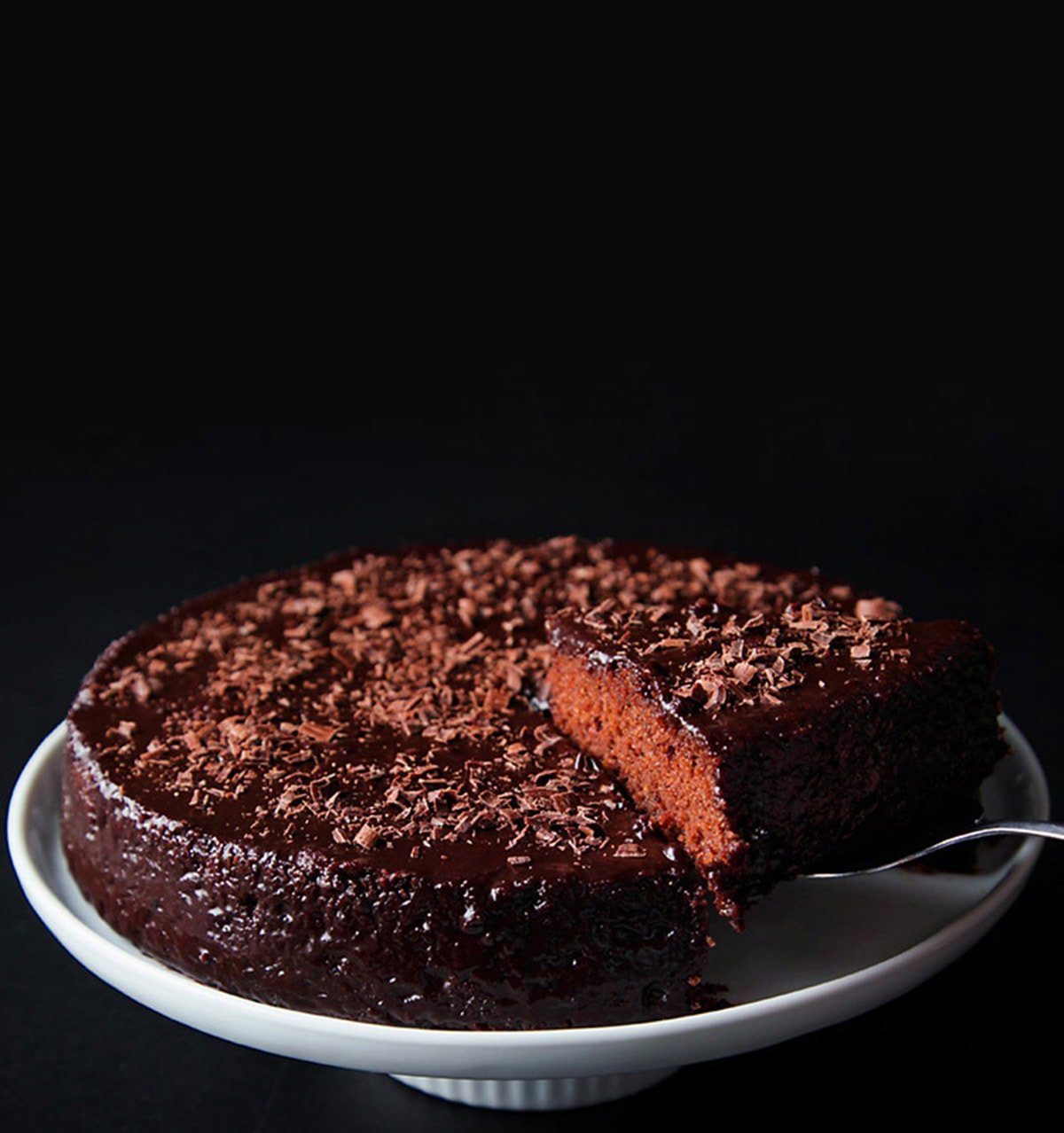 a slice of eggless chocolate cake being lifted with a spatula. The entire cake is kept on a white cake stand on a black board