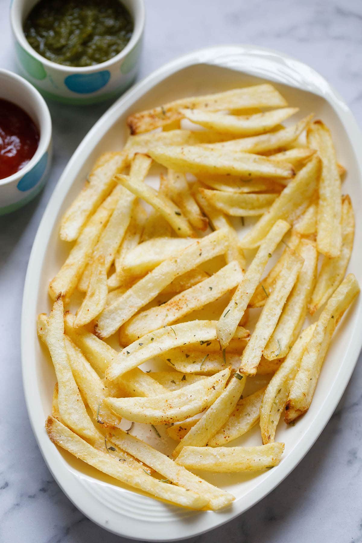 white serving tray filled with homemade french fries next to small dipping bowls of ketchup and green chutney