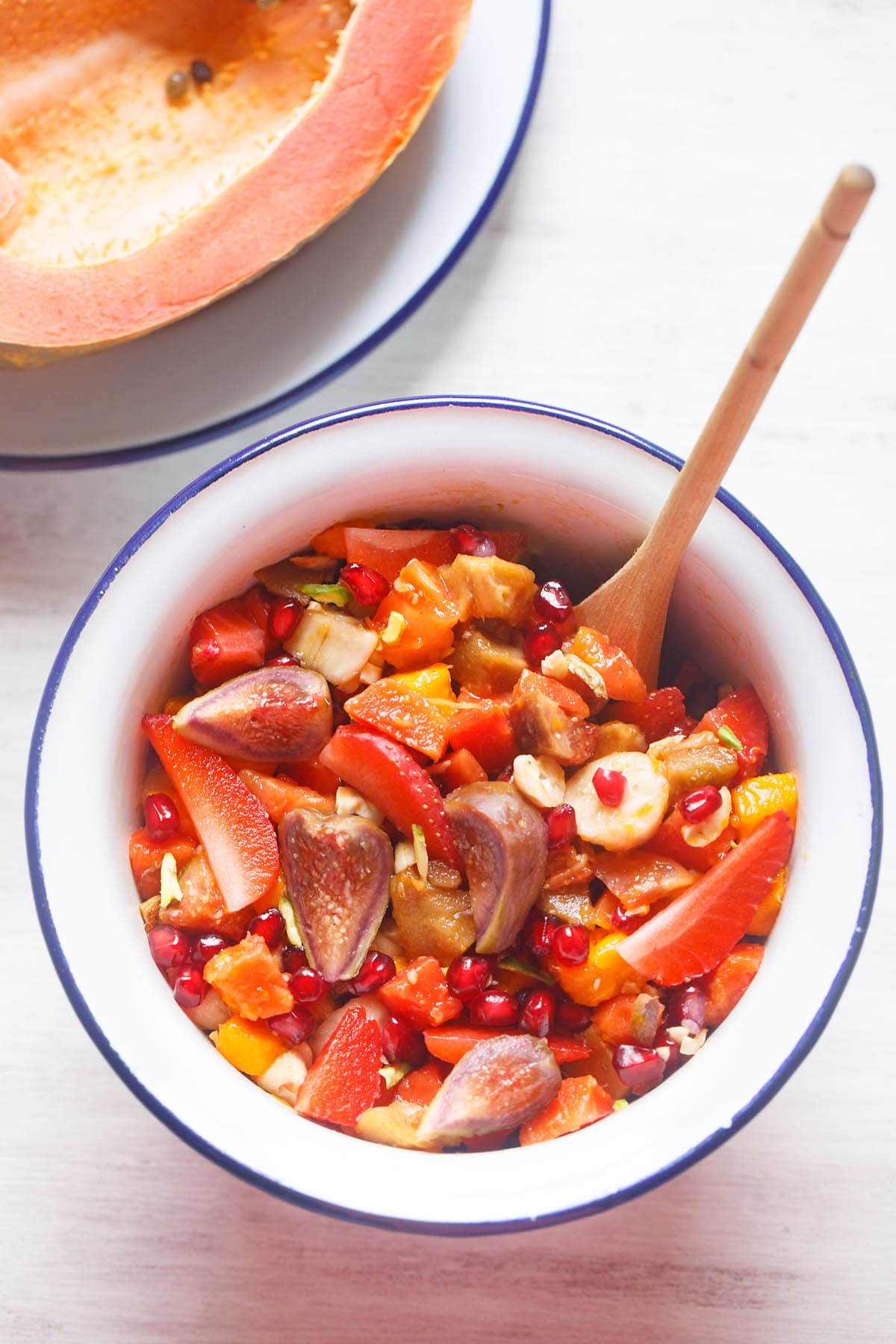 figs, strawberries, bananas, mango, chikoo, papaya and pomegranate tropical fruit salad in a white bowl next to a halved papaya