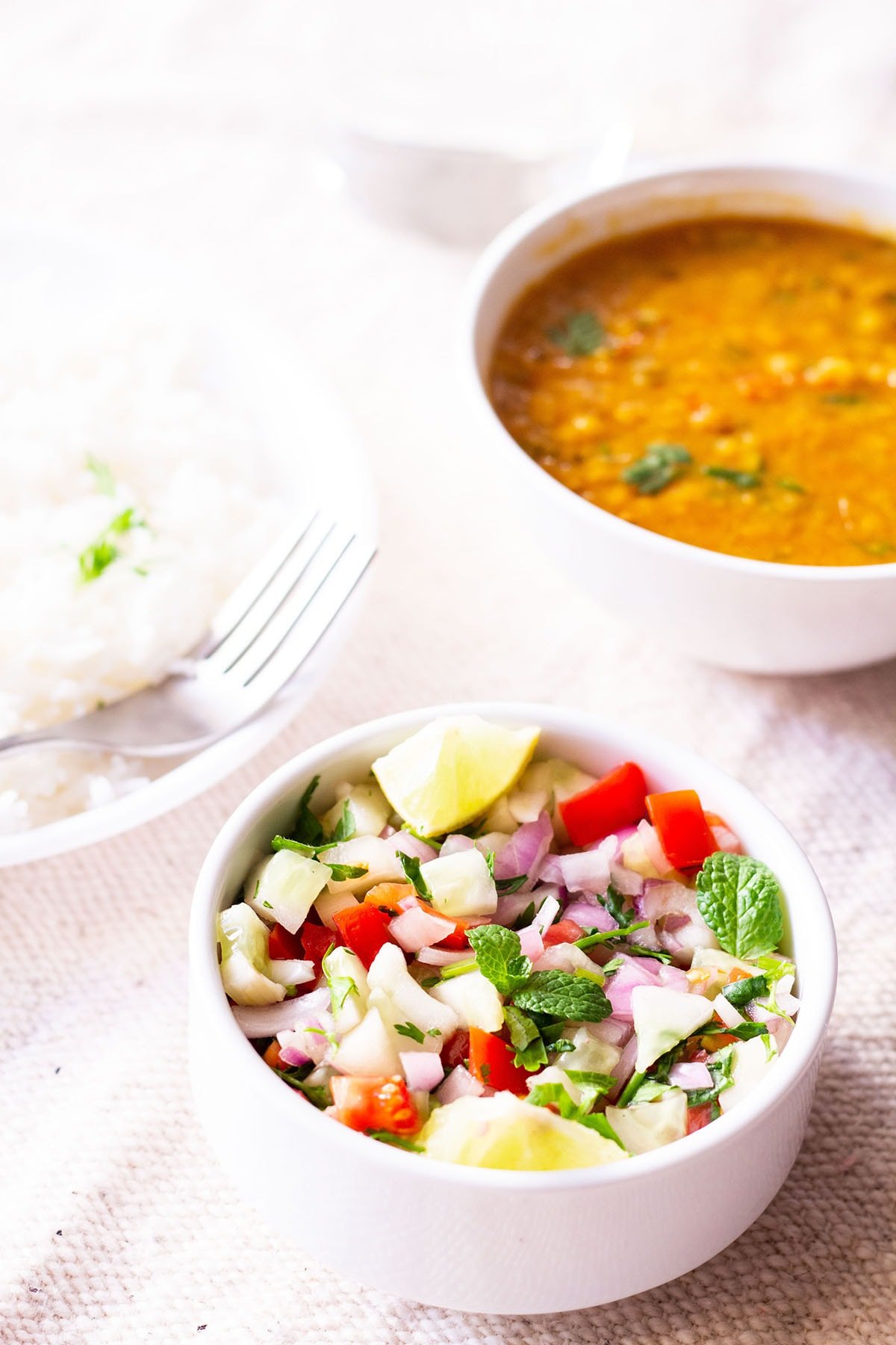 kachumber salad in a small white bowl on a table next to plate of rice and a bowl of lentils