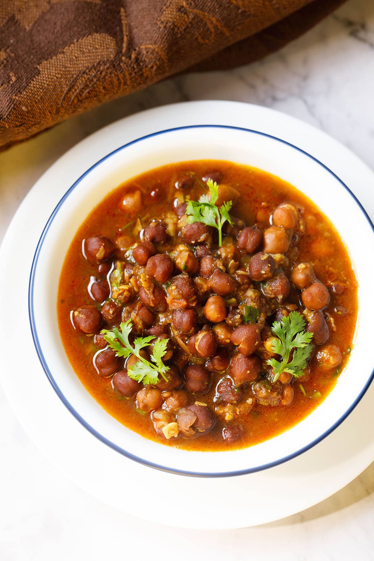 overhead shot of kala chana gravy with three coriander sprigs on top in a blue rimmed white bowl