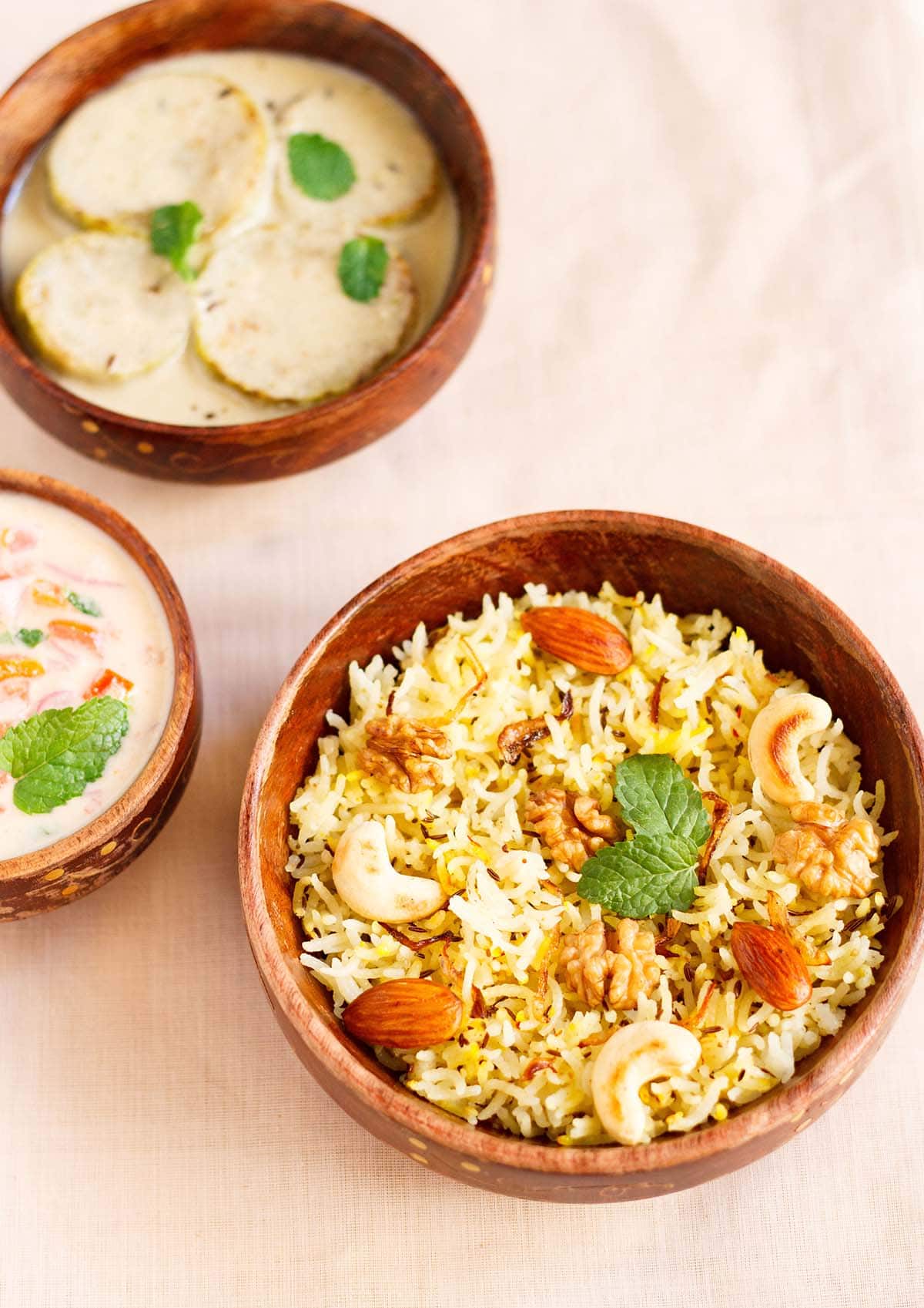 overhead shot of Kashmiri pulao in a brown bowl garnished with fried nuts and onions and a few fresh mint leaves next to a bowl of tomato raita on a beige table cloth
