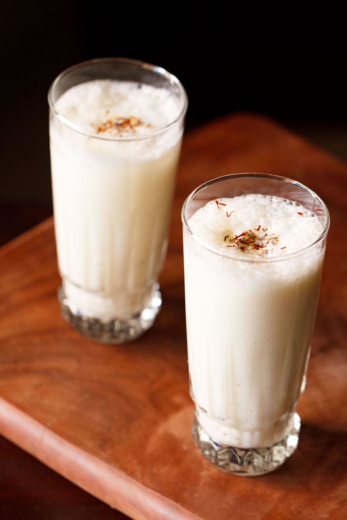 sweet lassi topped with crushed cardamom powder and saffron in two tall glasses on a wooden board