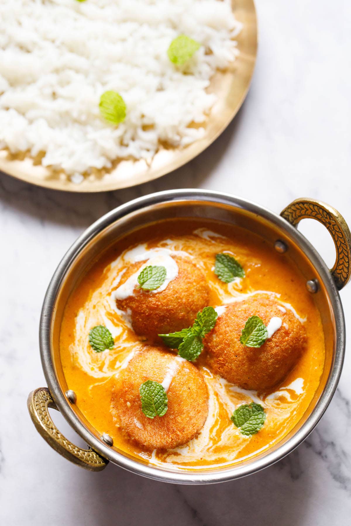 top shot of 3 malai kofta with mint leaves on top in a silver serving dish with handles on a white marble