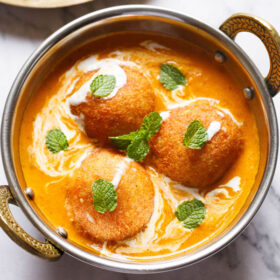 top shot of 3 malai kofta garnished with mint leaves in a silver serving dish with handles on a white marble
